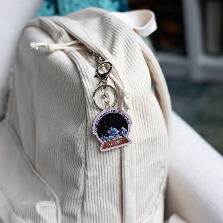  Acrylic keychain of snow globe with mountains against the night sky clipped to a white backpack. 'Court of Dreams City of Starlight' above it. ‘Velaris’ on the base. 

