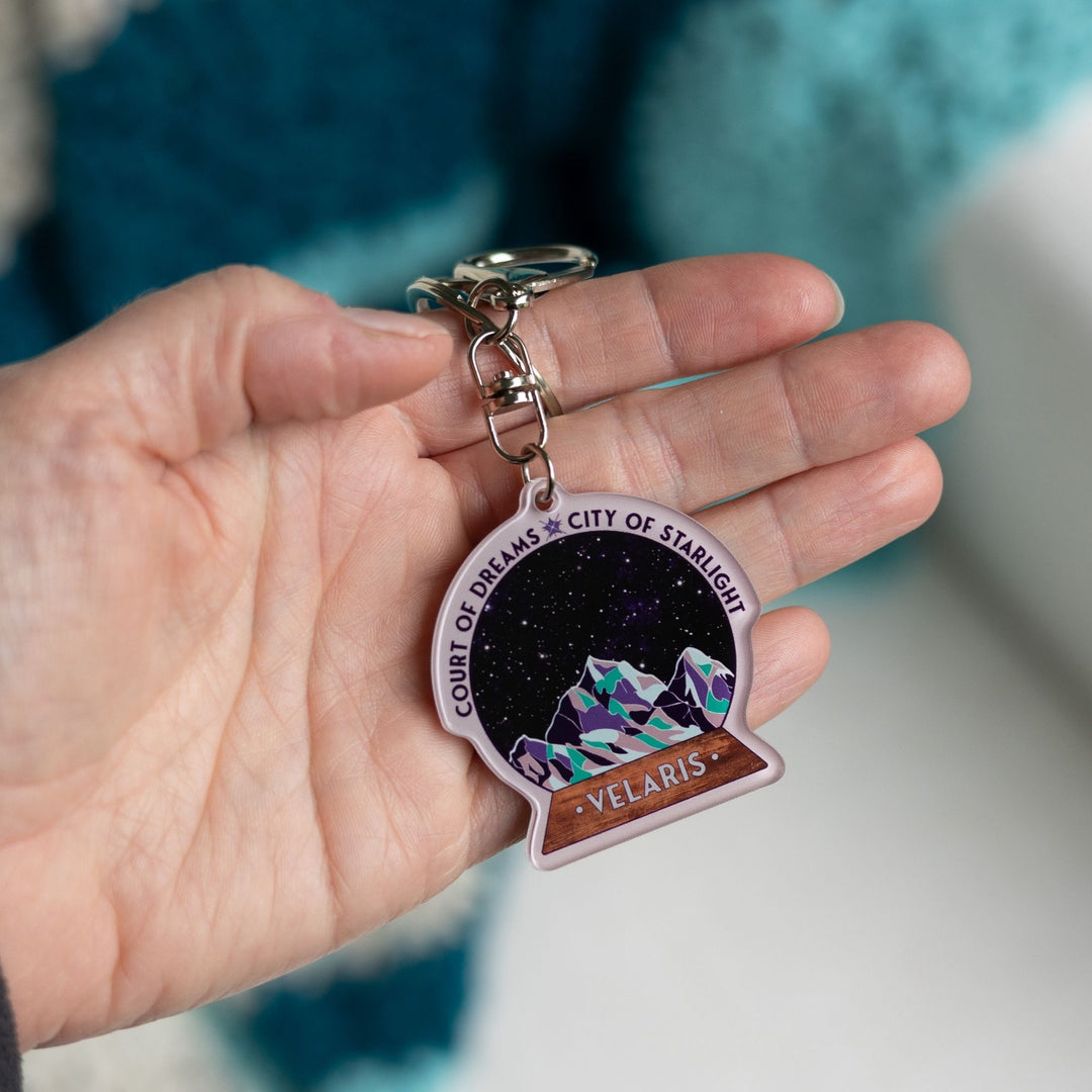 Acrylic keychain of snow globe with mountains against the night sky. 'Court of Dreams City of Starlight' above it. ‘Velaris’ on the base, held on a palm.
