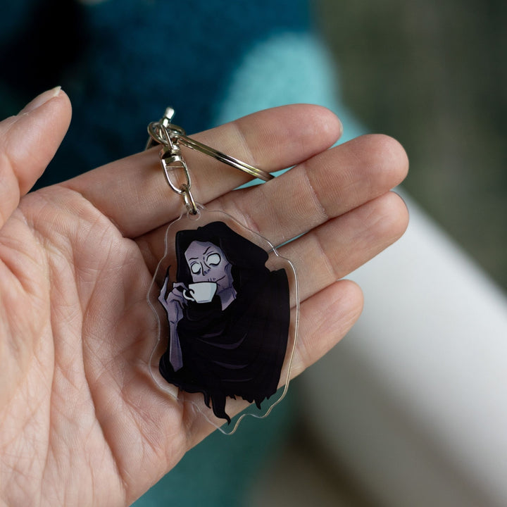 Acrylic keychain of the Suriel from A Court of Thorns and Roses, wearing a hood and sipping from a white tea cup, little finger raised, held on a palm.