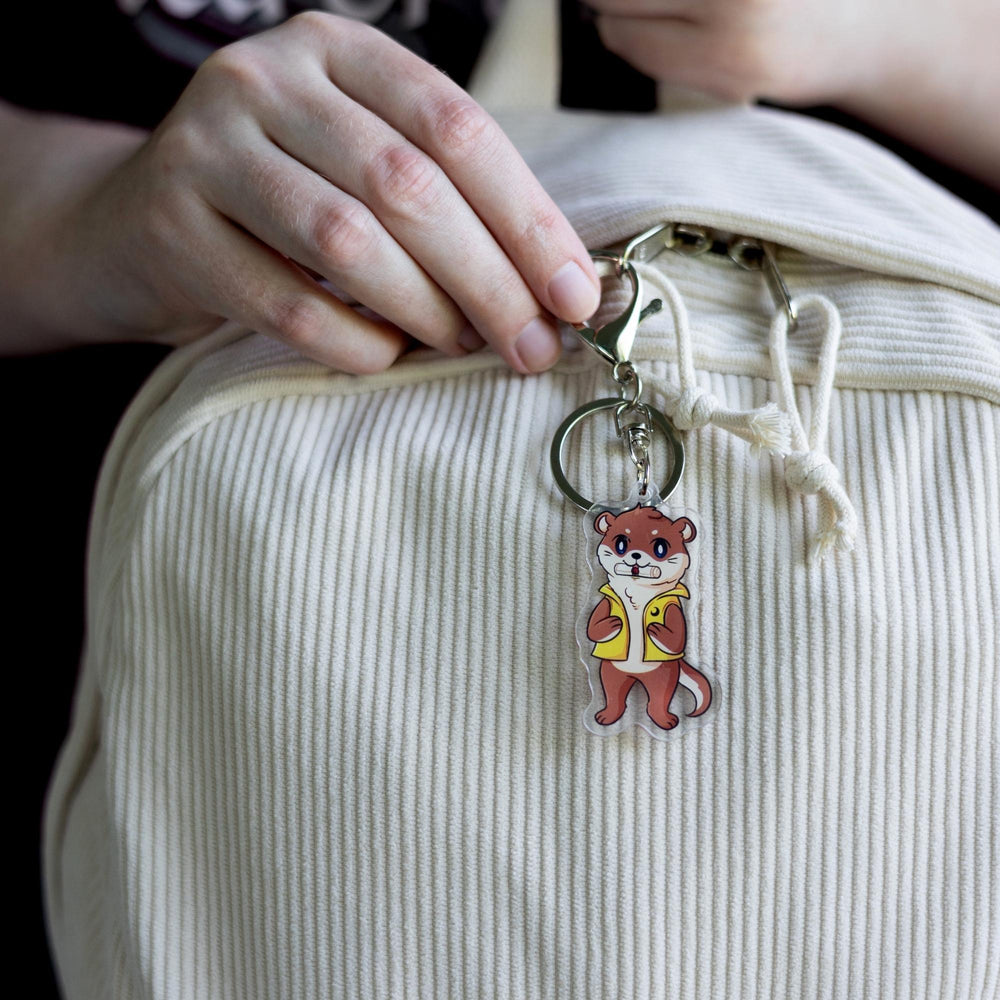Acrylic keychain of an otter with a scroll in its mouth, standing on its hind legs and wearing a yellow vest,  clipped to a white backpack.

