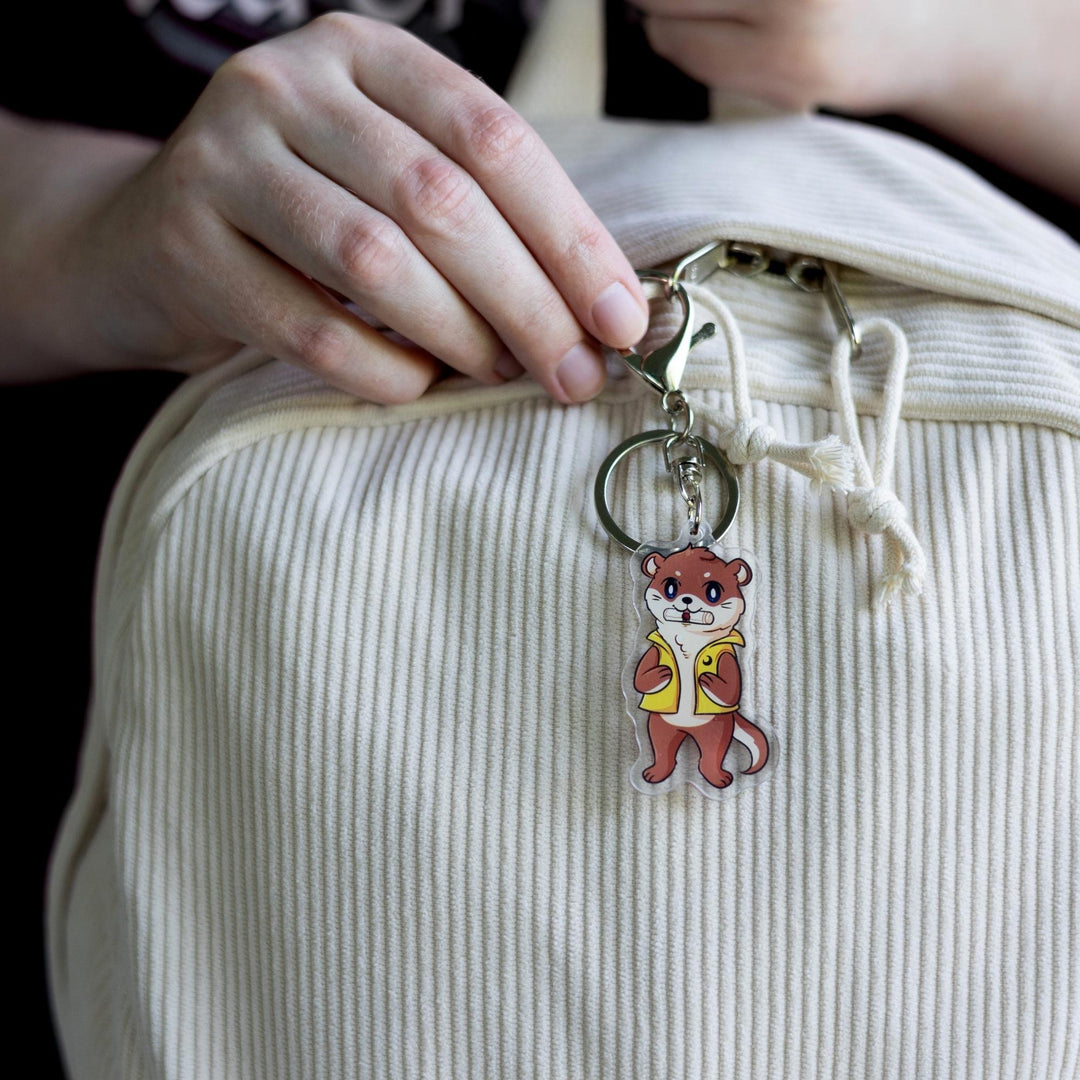 Acrylic keychain of an otter with a scroll in its mouth, standing on its hind legs and wearing a yellow vest,  clipped to a white backpack.
