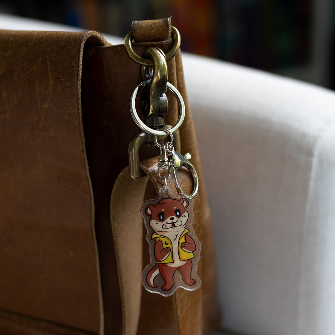  Acrylic keychain of an otter with a scroll in its mouth, standing on its hind legs and wearing a yellow vest, clipped to a leather purse. 
