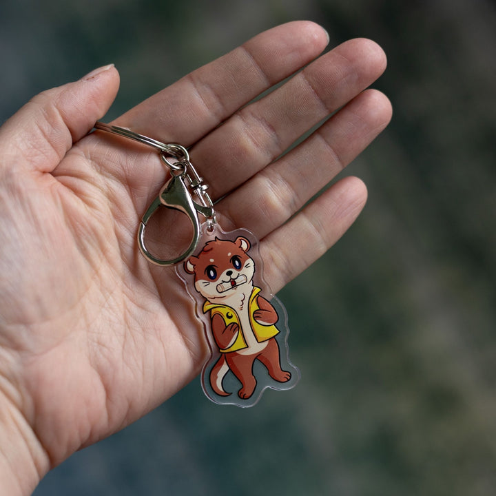  Acrylic keychain of an otter with a scroll in its mouth, standing on its hind legs and wearing a yellow vest, held on a palm.
