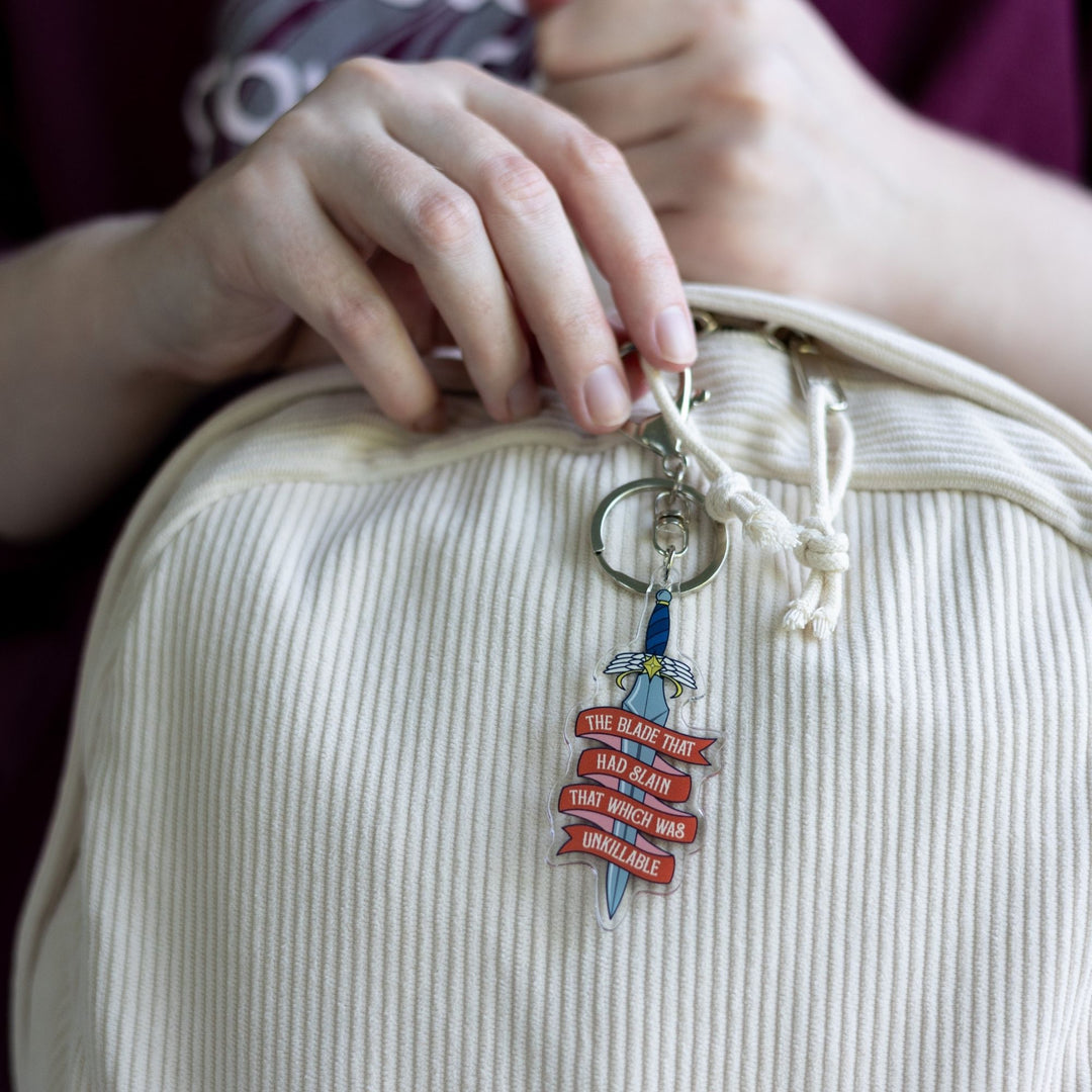  Acrylic keychain of a dagger clipped to a white backpack. On a red ribbon banner winding around the blade is ‘The blade that had slain that which was unkillable’ 
