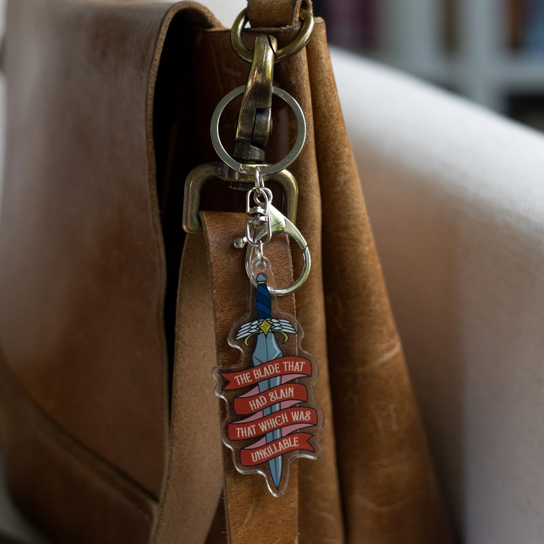 Acrylic keychain of a dagger clipped to a leather purse. On a red ribbon banner winding around the blade is ‘The blade that had slain that which was unkillable’ 