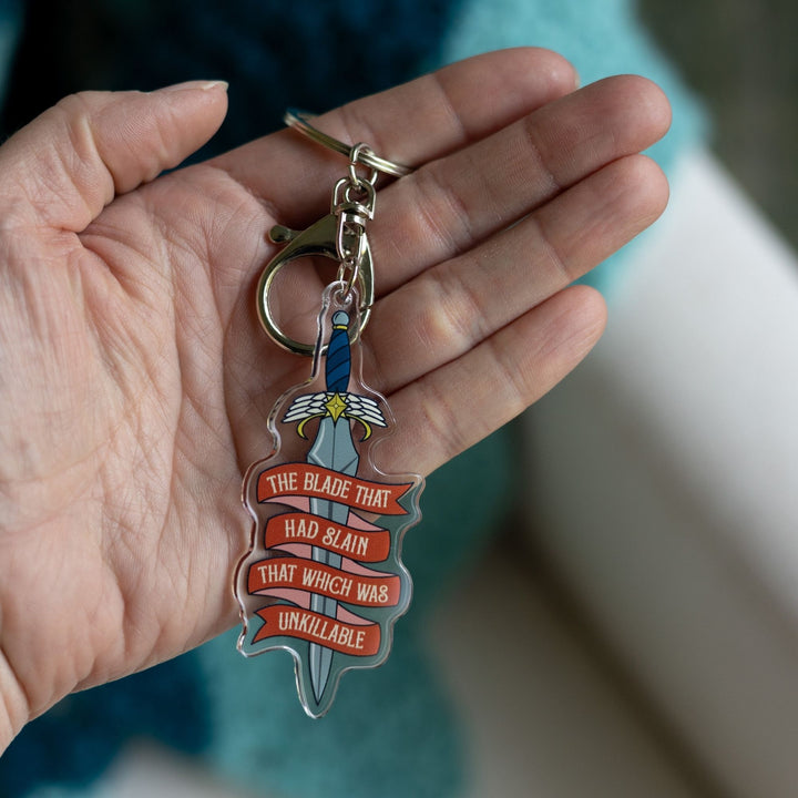 Acrylic keychain of a dagger held on a palm. On a red ribbon banner winding around the blade is ‘The blade that had slain that which was unkillable.’
