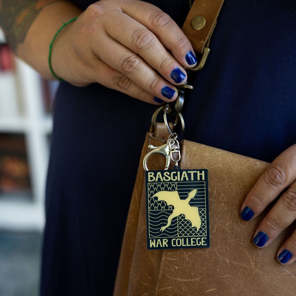 crylic keychain of black & gold Basgiath emblem, featuring a gold dragon flying over the quadrant symbols on a black background, clipped to a leather purse.