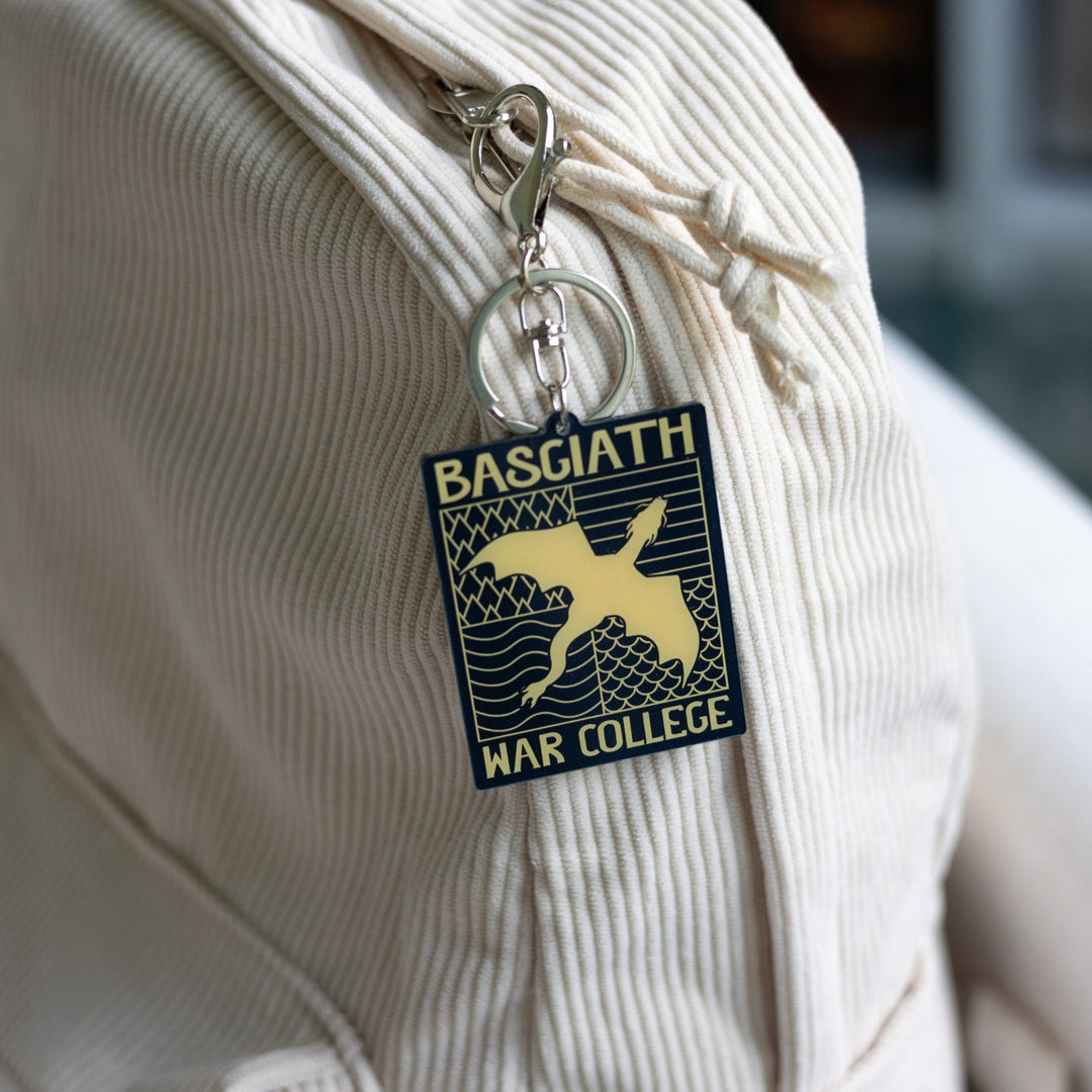 Acrylic keychain of black & gold Basgiath emblem, featuring a gold dragon flying over the quadrant symbols on a black background, clipped to a white backpack.
