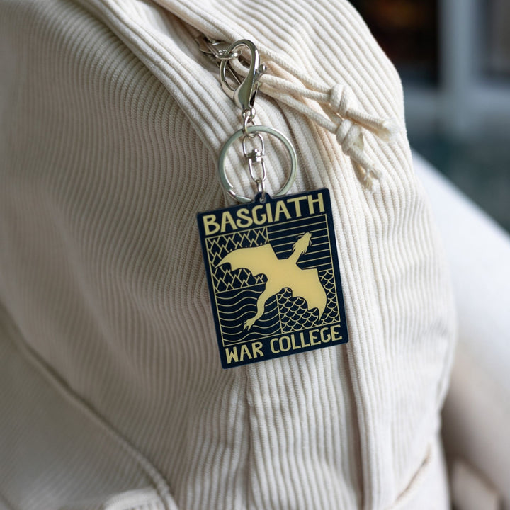 Acrylic keychain of black & gold Basgiath emblem, featuring a gold dragon flying over the quadrant symbols on a black background, clipped to a white backpack.
