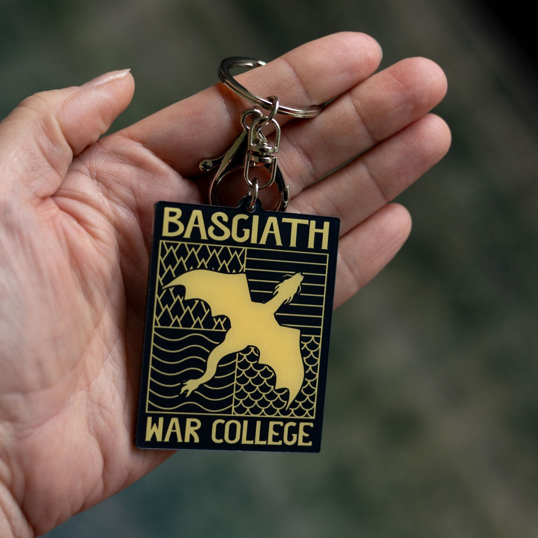 Acrylic keychain of black & gold Basgiath emblem, featuring a gold dragon flying over the quadrant symbols on a black background, held on a palm.
