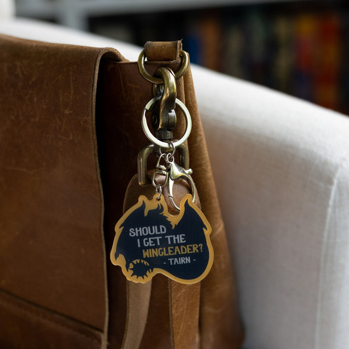 Acrylic keychain in the shape of a sitting dragon, black silhouette, with ‘Should I get the wingleader? Tairn’ in white text on it, clipped to a leather purse.
