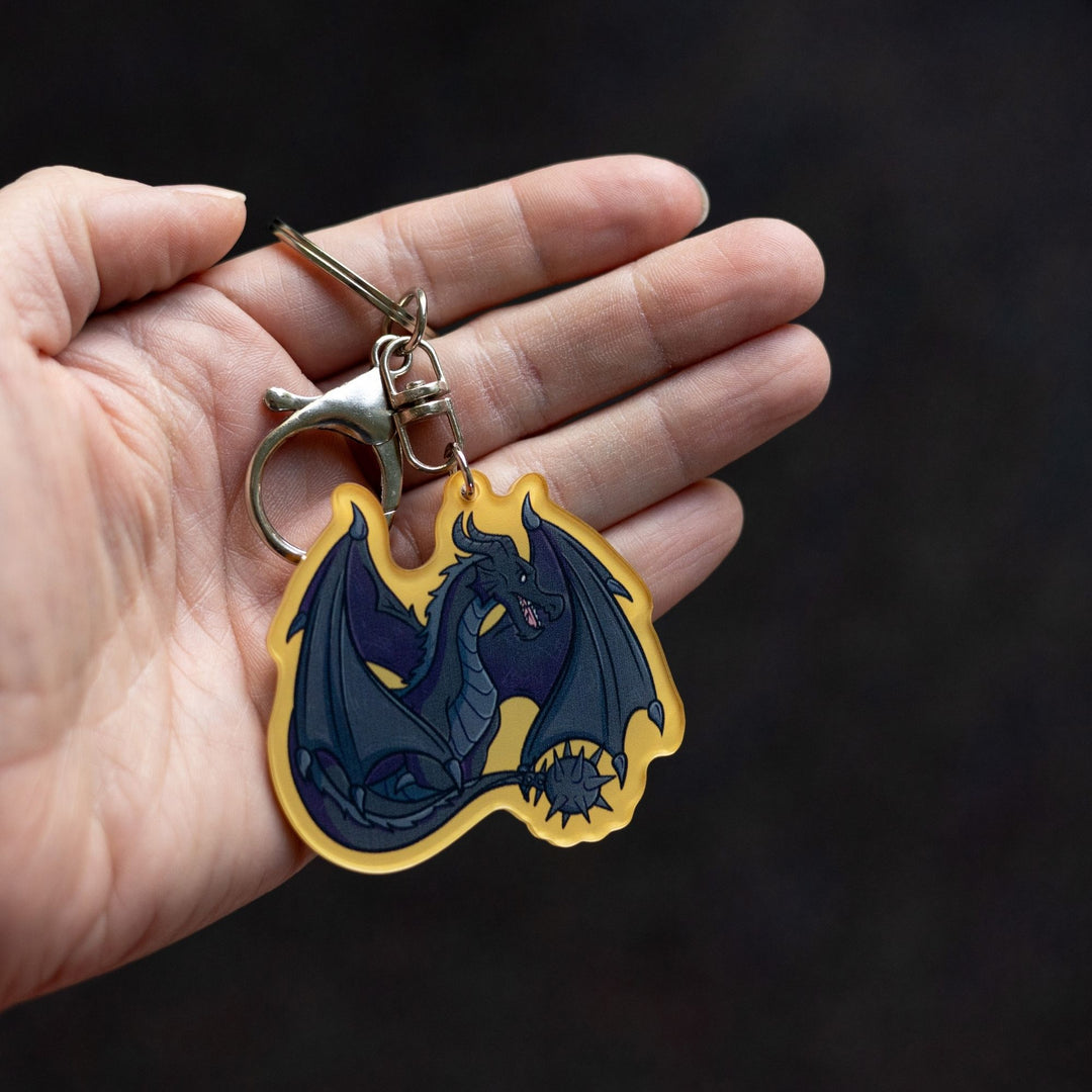 Acrylic keychain of Tairn, black dragon from Fourth Wing, sitting with tail and wings tucked around him with a golden yellow border, held on a palm.
