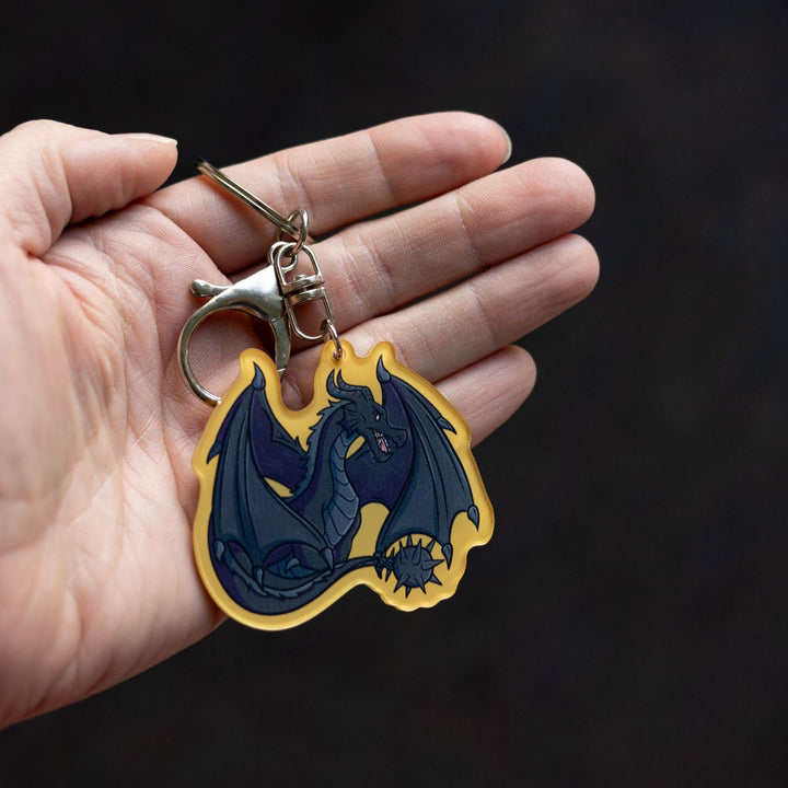 Acrylic keychain of Tairn, black dragon from Fourth Wing, sitting with tail and wings tucked around him with a golden yellow border, held on a palm.