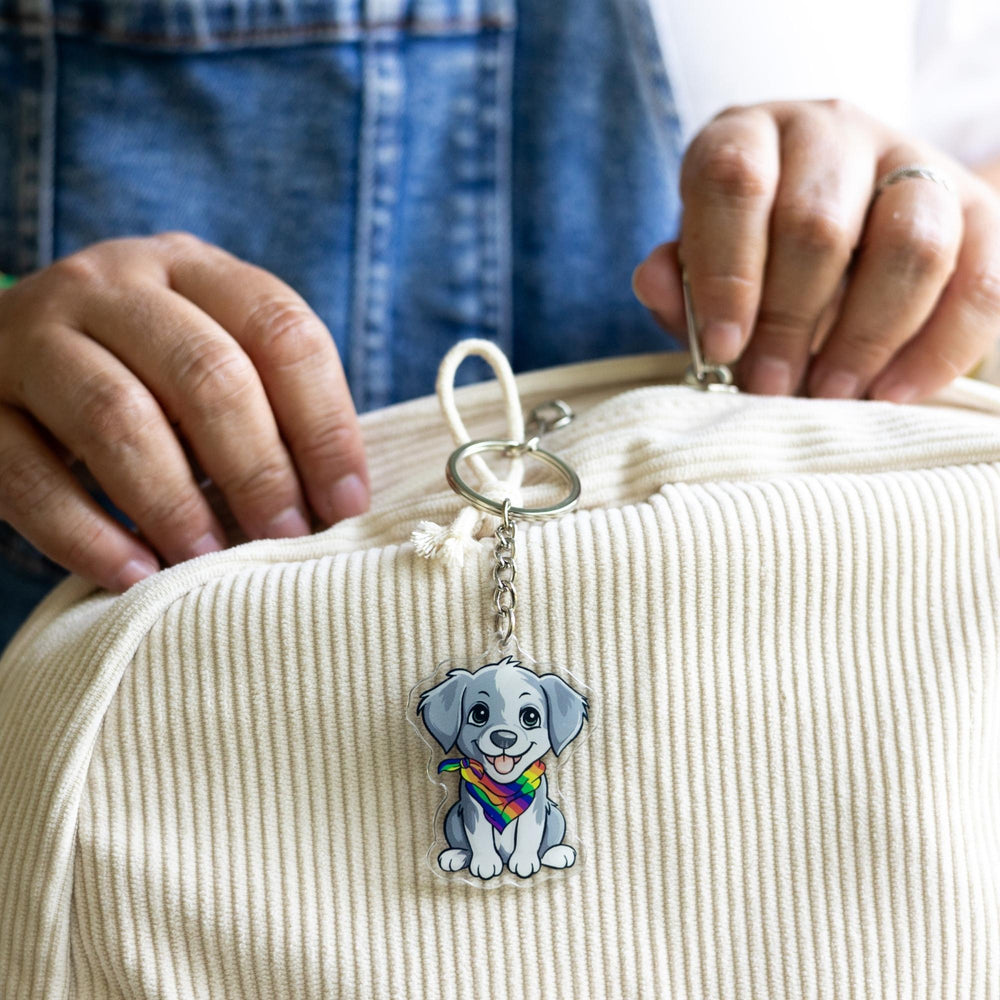  Acrylic keychain of a grey and white puppy, sitting happily and wearing a rainbow pride bandana clipped to a white backpack.

