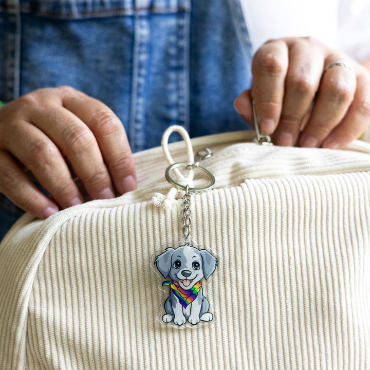  Acrylic keychain of a grey and white puppy, sitting happily and wearing a rainbow pride bandana clipped to a white backpack.
