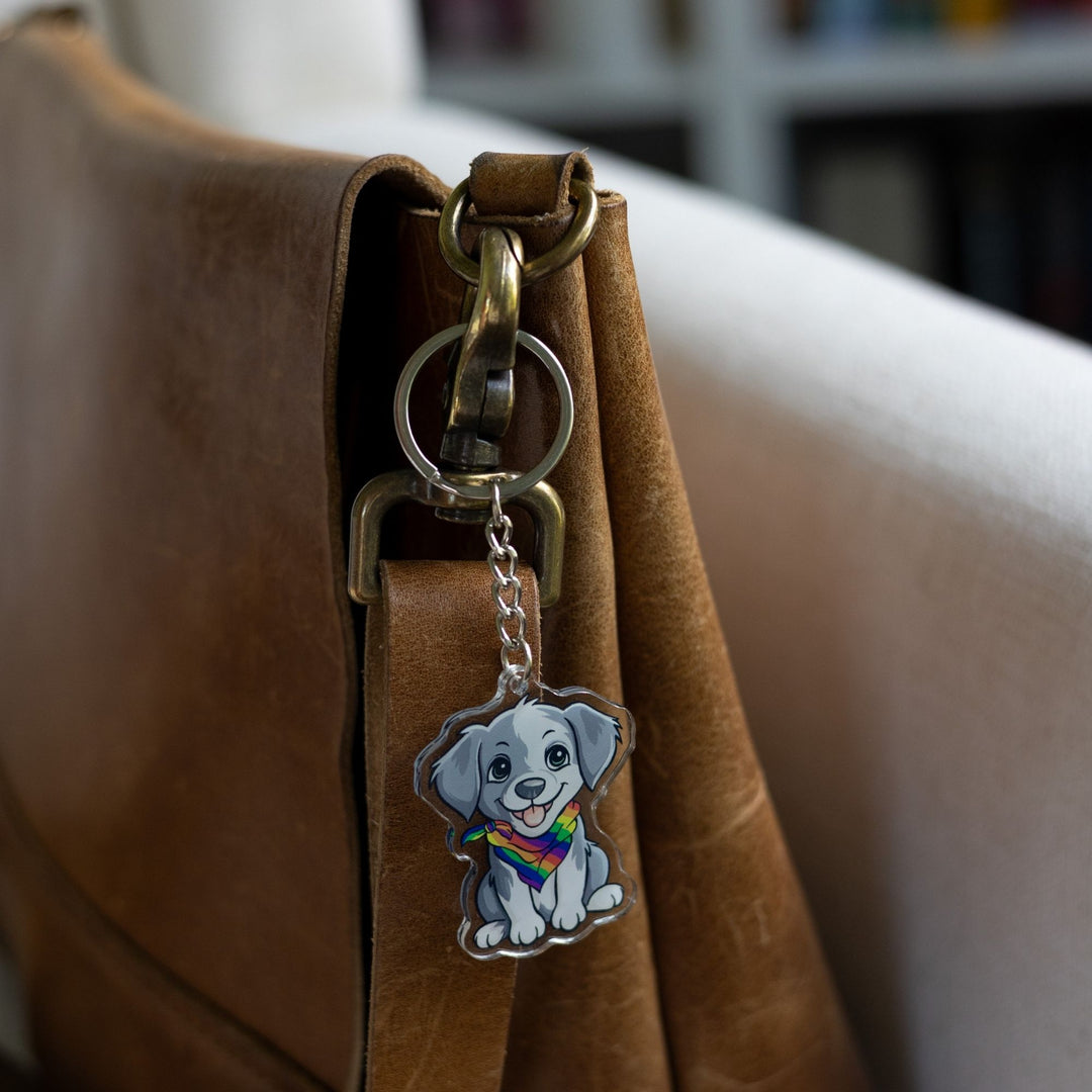 : Acrylic keychain of a grey and white puppy, sitting happily and wearing a rainbow pride bandana clipped to a leather purse.
