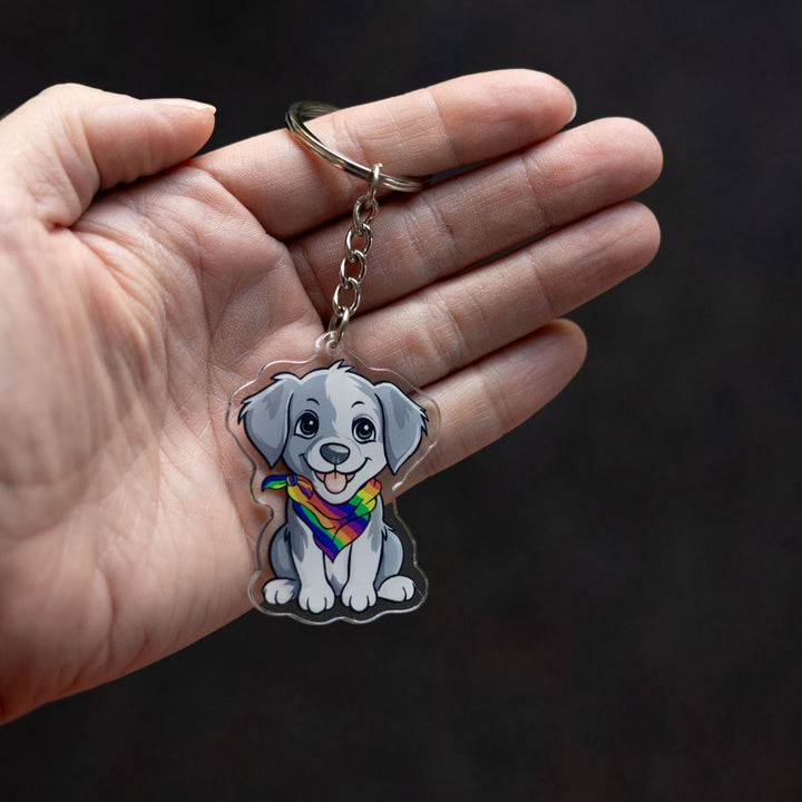 Acrylic keychain of a grey and white puppy, sitting happily and wearing a rainbow pride bandana held in a palm.