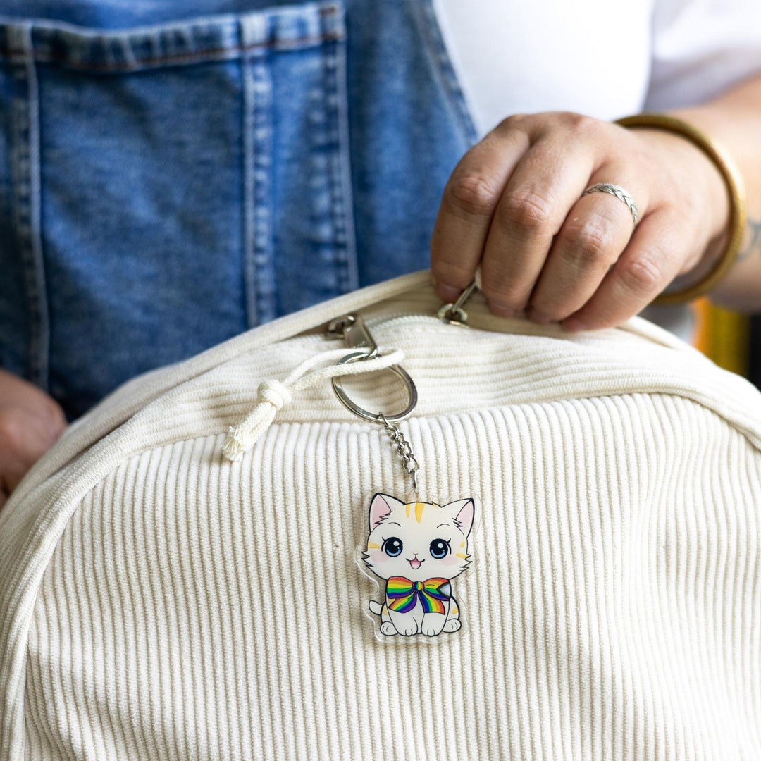  Acrylic keychain of a cream colored kitten with orange stripes, sitting happily and wearing a progress pride flag bow tie  clipped to a white backpack.
