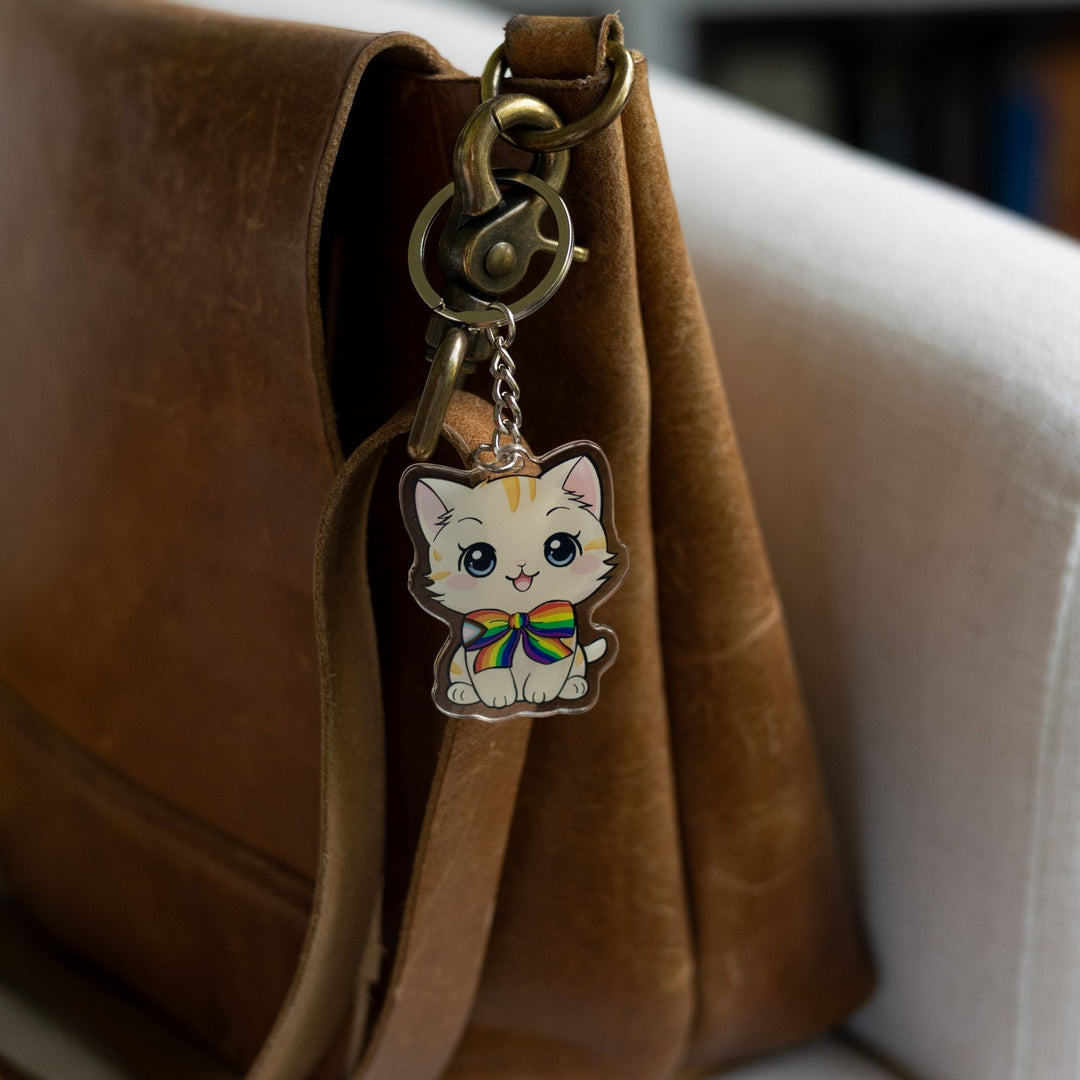 Acrylic keychain of a cream colored kitten with orange stripes, sitting happily and wearing a progress pride flag bow tie clipped to a leather purse.
