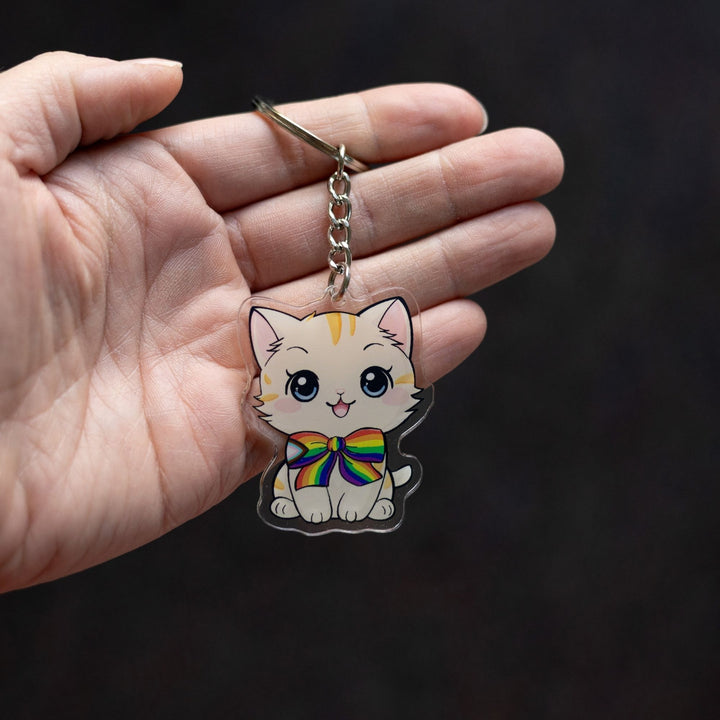  Acrylic keychain of a cream colored kitten with orange stripes, sitting happily and wearing a progress pride flag bow tie, held in a palm