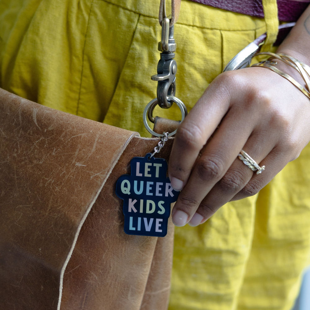 Acrylic keychain that says ‘Let queer kids live.’ The words are a pastel rainbow on a black background clipped to a leather purse.
