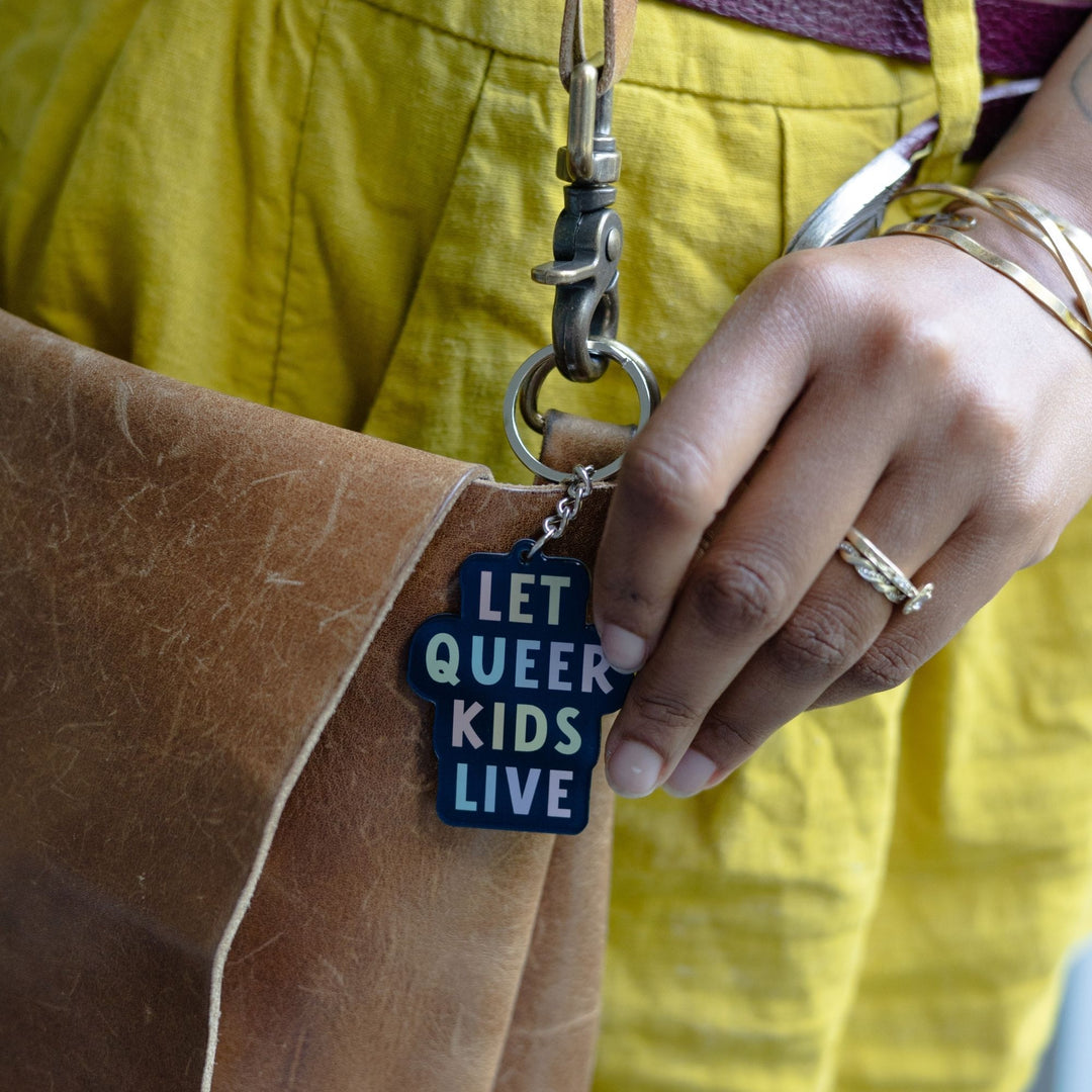 Acrylic keychain that says ‘Let queer kids live.’ The words are a pastel rainbow on a black background clipped to a leather purse.
