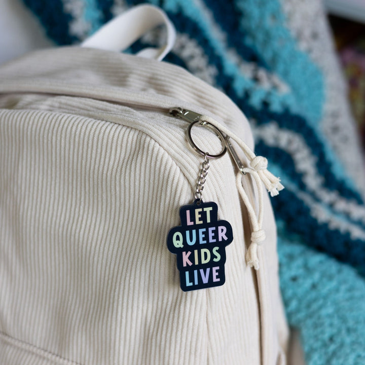 Acrylic keychain that says ‘Let queer kids live.’ The words are a pastel rainbow on a black background clipped to a white backpack.