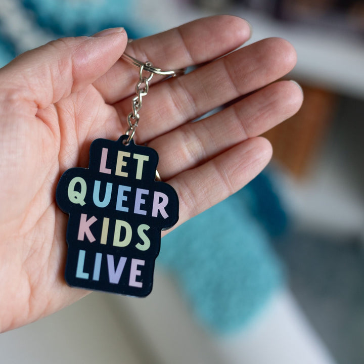  Acrylic keychain that says ‘Let queer kids live.’ The words are a pastel rainbow on a black background, held on a palm.

