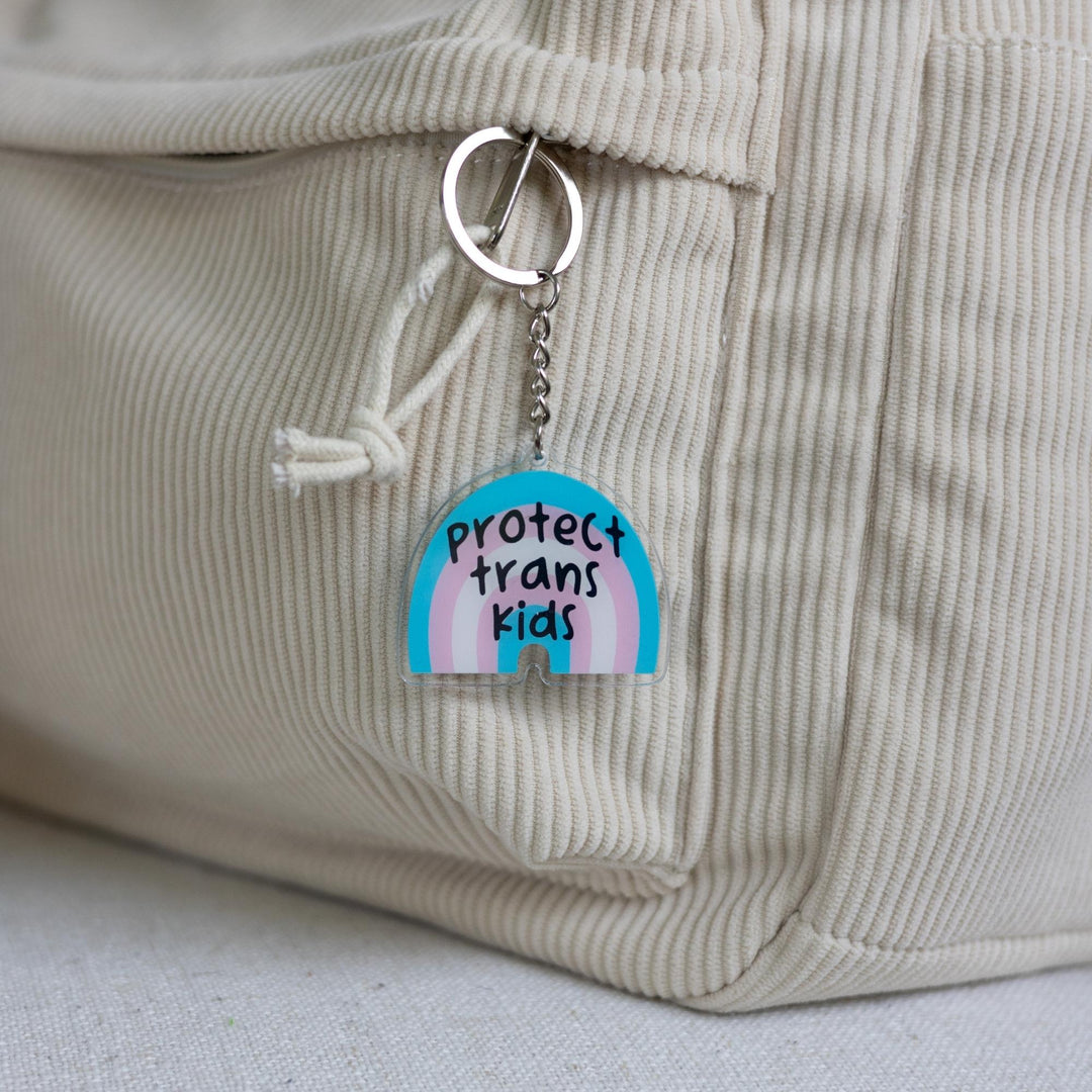 Acrylic keychain of a trans pride rainbow that says ‘Protect trans kids.’ The rainbow is blue, pink, and white clipped to a white backpack.
