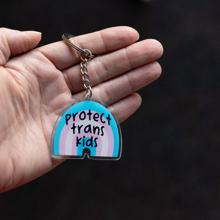 Acrylic keychain of a trans pride rainbow that says ‘Protect trans kids.’ The rainbow is blue, pink, and white, held on a palm.

