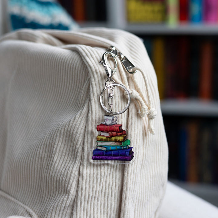 Acrylic keychain of a stack of books that are each a different color of the rainbow with a tea cup with a pink flower and saucer on top  clipped to a white backpack.
