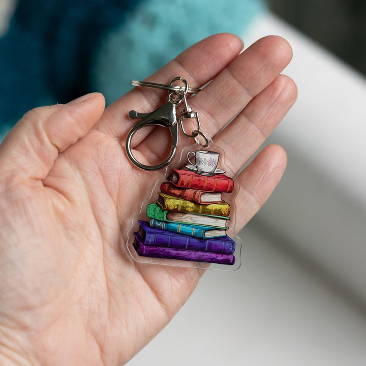Acrylic keychain of a stack of books that are each a different color of the rainbow with a tea cup with a pink flower and saucer on top held in a palm
