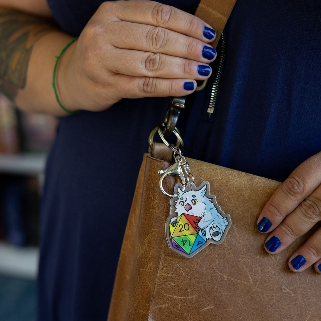  Acrylic keychain of a baby Owlbear holding a rainbow d20 clipped to a leather purse. The number 20 on the die is facing up.
