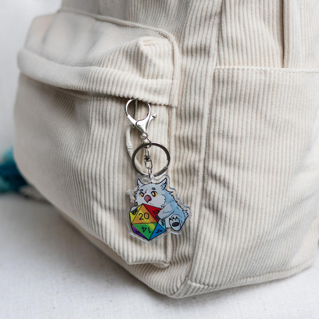  Acrylic keychain of a baby Owlbear holding a rainbow d20 clipped to a white backpack. The number 20 on the die is facing up.
