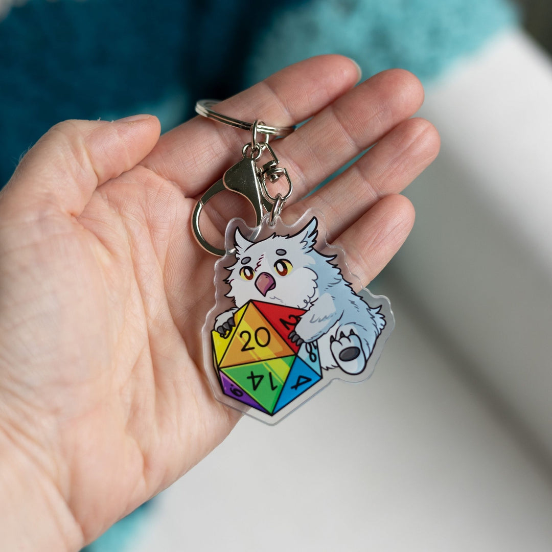 Acrylic keychain of a baby Owlbear holding a rainbow d20 held in a palm. The number 20 on the die is facing up.
