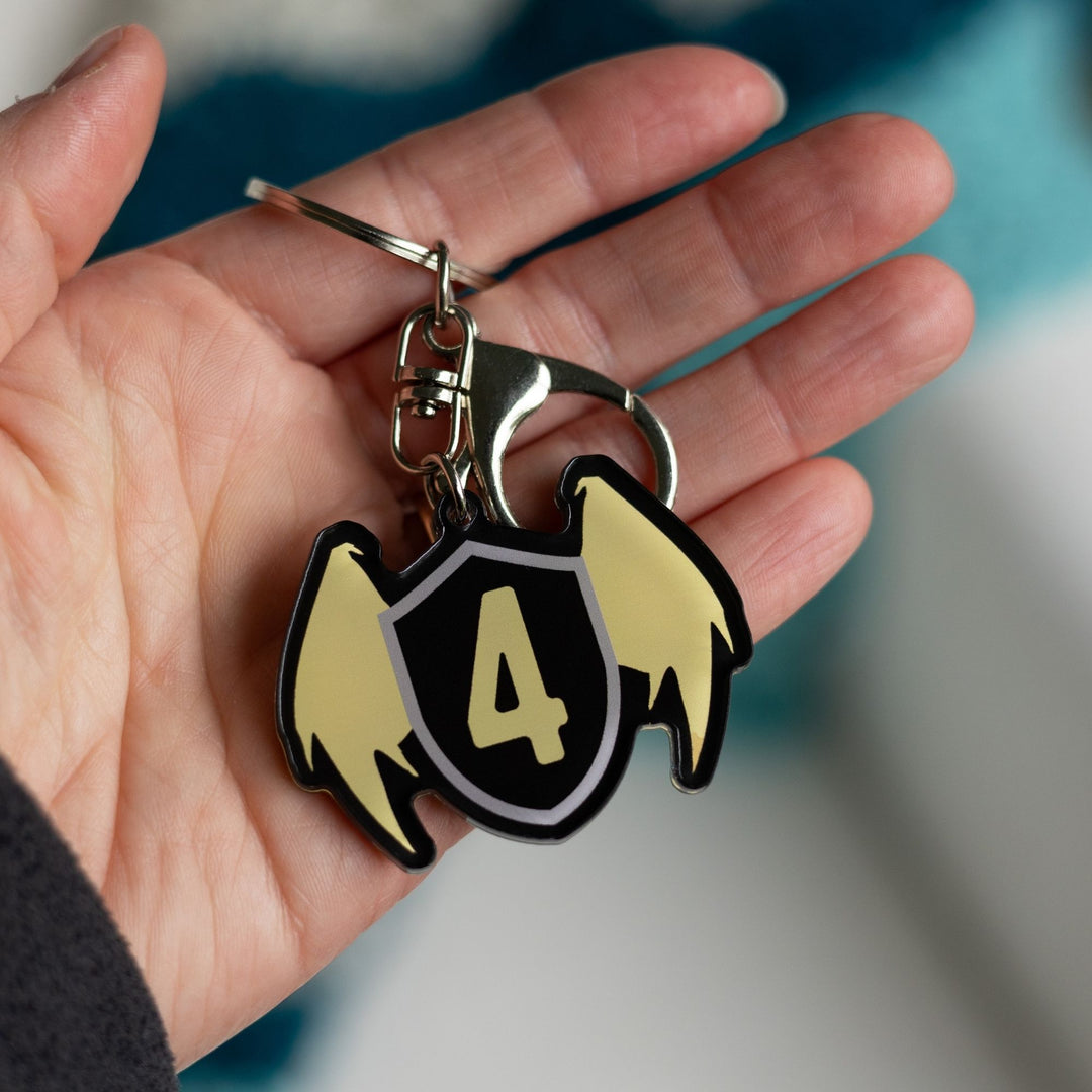 Acrylic keychain of a black shield with golden wings & a gold four on the shield held in a palm. From Fourth Wing by Rebecca Yarros.
