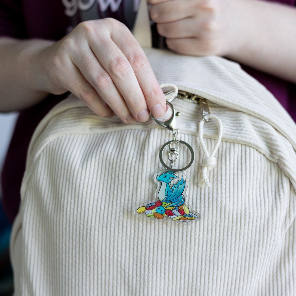 Acrylic keychain of Theodore, a blue baby  wyvern, sitting on a pile of multicolored buttons,  clipped to a white backpack.
