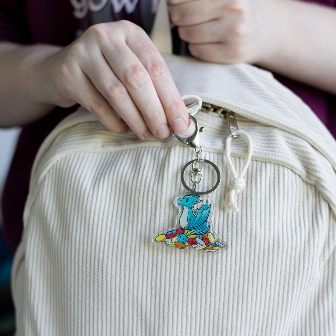 Acrylic keychain of Theodore, a blue baby  wyvern, sitting on a pile of multicolored buttons,  clipped to a white backpack.
