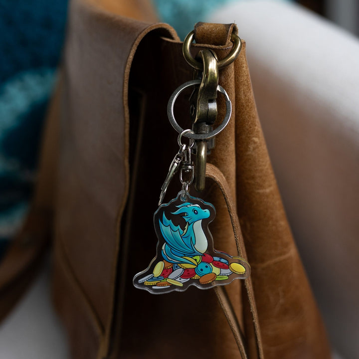 Acrylic keychain of Theodore, a blue baby wyvern, sitting on a pile of multicolored buttons clipped to a leather purse.

