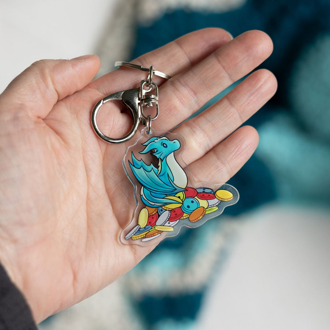  Acrylic keychain of Theodore, a blue baby wyvern, sitting on a pile of multicolored buttons held in a palm. From House in the Cerulean Sea.
