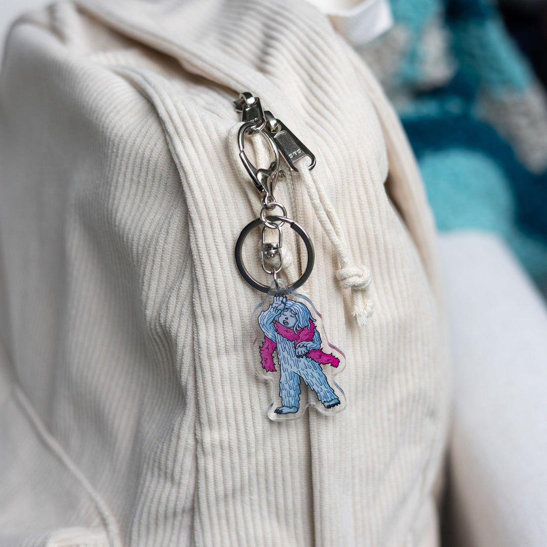 : Acrylic keychain of David the Yeti wearing a pink boa and hand dramatically against their head a clipped to a white backpack.
