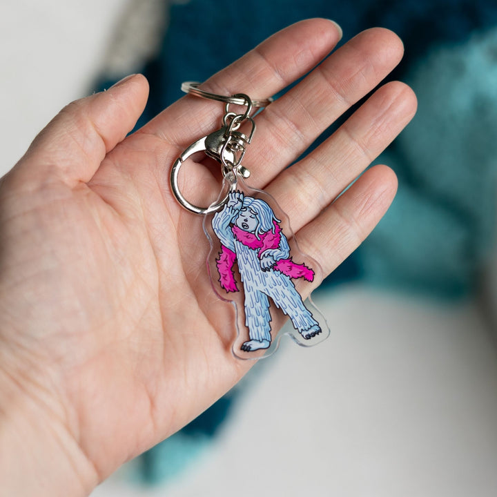 crylic keychain of David the Yeti wearing a pink boa and hand dramatically against their head held in a palm. From House in the Cerulean Sea.
