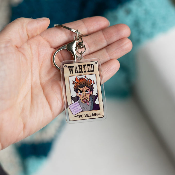 Acrylic keychain of a wanted poster of ‘The Villain’. With a purple post-it note on it that reads ‘An Uncanny resemblance, Your evilness’ held in a palm