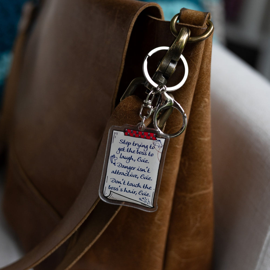  Acrylic keychain of a scroll with a list: ‘Stop trying to get the boss to laugh, Evie. Danger isn’t attractive, Evie. Don’t touch the boss’s hair, Evie’ clipped to a leather purse.
