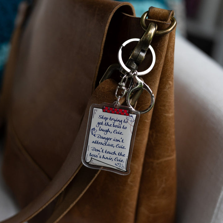  Acrylic keychain of a scroll with a list: ‘Stop trying to get the boss to laugh, Evie. Danger isn’t attractive, Evie. Don’t touch the boss’s hair, Evie’ clipped to a leather purse.
