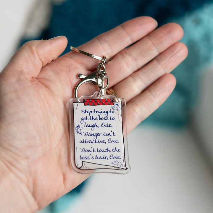 Acrylic keychain of a scroll with a list: ‘Stop trying to get the boss to laugh, Evie. Danger isn’t attractive, Evie. Don’t touch the boss’s hair, Evie’ held in a palm.