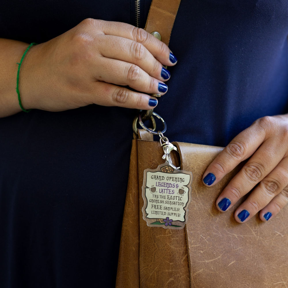 Acrylic keychain of parchment sign nailed to a post: ‘Grand opening. Legends & Lattes. Try the exotic gnomish sensation! Free samples! Limited Supply.' clipped to a leather purse.
