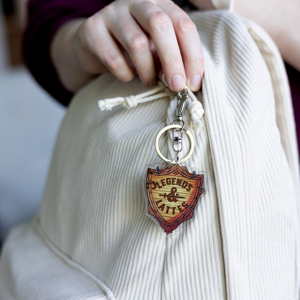Acrylic keychain of a wooden shield with ‘Legends & Lattes’ shop logo on it clipped to a white backpack