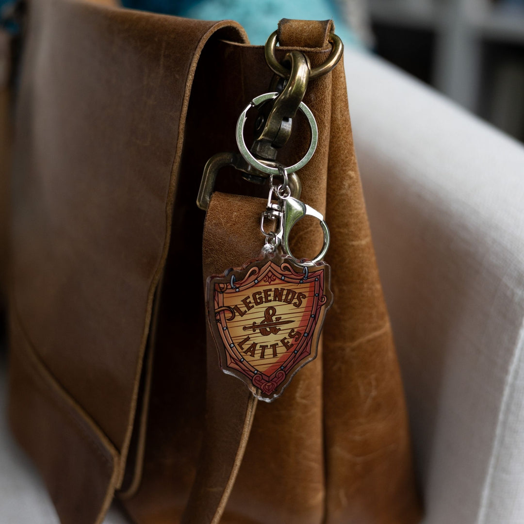   Acrylic keychain of a wooden shield with ‘Legends & Lattes’ shop logo on it clipped to a clipped to a leather purse.
