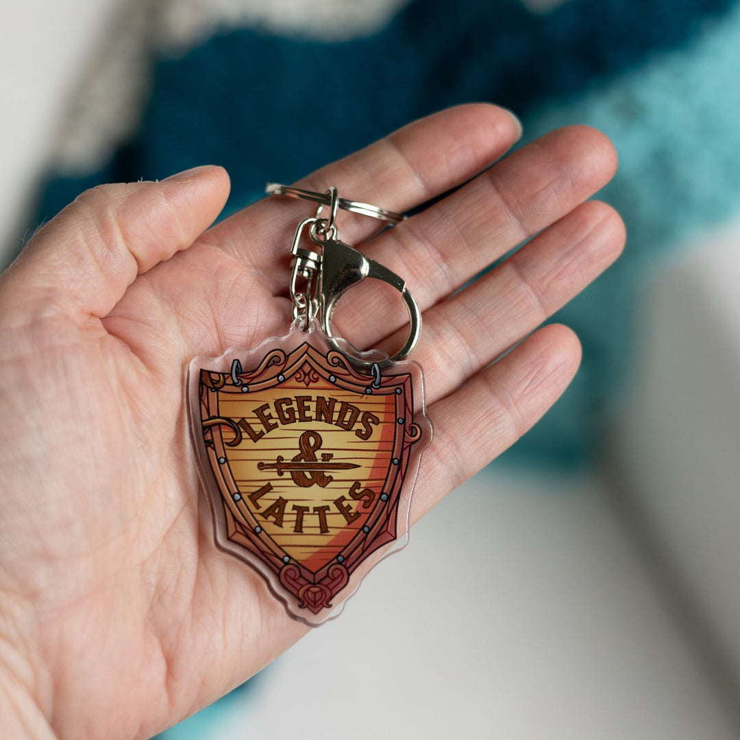 Acrylic keychain of a wooden shield with ‘Legends & Lattes’ shop logo on it held in a palm.
